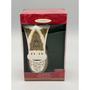 May include: A Hallmark Keepsake ornament of a Nutcracker figure, designed by Tracy Larsen. The ornament is white with gold accents and a detailed design. The packaging is red and includes the text "Hallmark Keepsake Ornament".