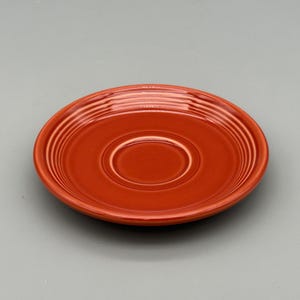 May include: A round, orange-red ceramic saucer with concentric rings. The saucer has a slightly recessed center and a glossy finish. This vintage-style saucer is suitable for a teacup or coffee cup.