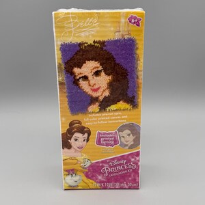 May include: A Disney Princess Belle latch hook kit in a rectangular box. The packaging features a purple latch hook design of Belle, a yellow background, and text including Disney Princess Latch Hook Kit and 12 IN x 12 IN. It includes pre-cut yarn.