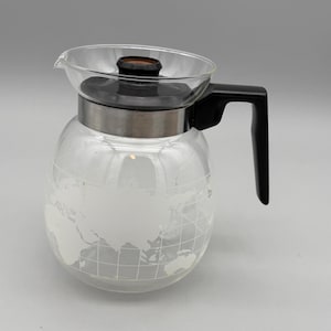 May include: A clear glass coffee carafe featuring a frosted white world map design etched onto the exterior. The carafe includes a black plastic lid with a central brown accent, a silver-toned metal band around the neck, and a black handle.