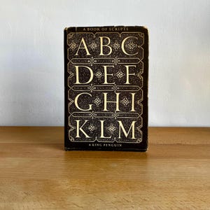 以下が含まれることがあります： 「A Book of Scripts」というタイトルのヴィンテージ本。暗い背景に金色の文字が書かれています。表紙には、装飾的なフォントでABC、DEF、GHI、KLMの文字が描かれています。本は木製の表面に置かれています。