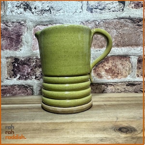 May include: A lime green ceramic pitcher with a speckled glaze finish. The vessel features a smooth upper section, a series of three horizontal raised ridges around the lower body, a curved handle on the right side, and a flat circular base.