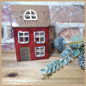 May include: A red ceramic house-shaped candle holder with a brown roof. The house has four square windows and a door. The windows and door are outlined in white. The candle holder is on a wooden surface.