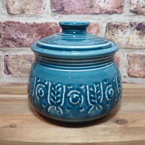 May include: A teal ceramic jar with a matching lid, featuring a rounded body and a textured band of embossed leaf and circular patterns around the centre. The jar has a glossy finish and horizontal ridges near the top rim and on the lid.