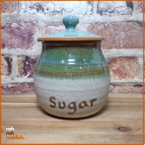 May include: A ceramic sugar canister with a lid, featuring a rounded body and a tapered neck. The upper portion is glazed in a glossy green, while the lower section is a speckled off-white. The word Sugar is inscribed in brown on the front.