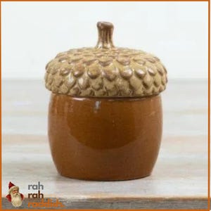 May include: A ceramic jar shaped like an acorn with a smooth, glossy brown base and a textured, tan-coloured cap featuring a small stem on top. The jar is decorated with the text rah rah roddish in the bottom left corner of the image.