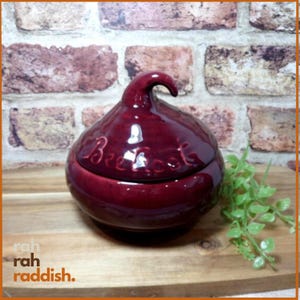 May include: A glossy, deep burgundy ceramic jar with a rounded body and a curved, stem-like lid. The word "Beetroot" is embossed in cursive script on the front of the jar. The ceramic surface has a smooth, reflective finish throughout.