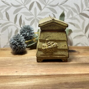May include: A ceramic beehive-shaped container with a lid, in a speckled yellow-green colour. The container has a detailed bee design on the front. The background includes a wooden surface and floral wallpaper.