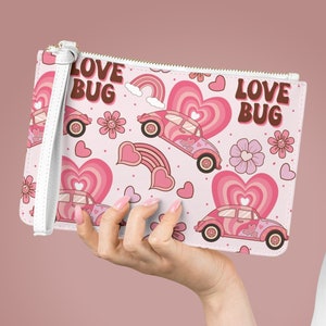 May include: A pink clutch with a repeating pattern of pink cars, hearts, rainbows, and flowers. The words "LOVE BUG" are printed on the top. The clutch has a white wrist strap and a gold zipper.