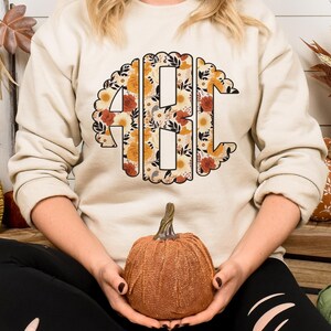 May include: A beige sweatshirt with a floral monogram design featuring the letters "ABC" in a circle. The sweatshirt is worn by a person sitting with a pumpkin in their lap.