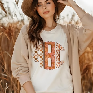 May include: A white t-shirt with a large, colorful monogram in the center. The monogram is made up of three letters, "ABC", each with a different pattern. The letters are surrounded by a scalloped border.