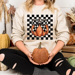 May include: A beige sweatshirt with a black and white checkered design featuring a pumpkin with a black and white bow. The pumpkin is orange with a green stem.