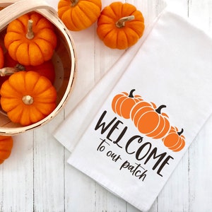May include: White kitchen towel with the text "WELCOME to our patch" in black lettering and a graphic of three orange pumpkins. A basket of orange pumpkins is in the background.