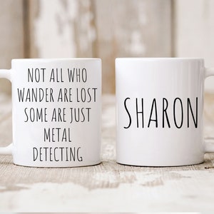 May include: Two white ceramic mugs. One mug has the text "Not all who wander are lost some are just metal detecting". The other mug has the name "SHARON" written on it.