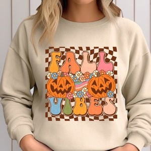 May include: A beige sweatshirt with a retro-style graphic design featuring the text "Fall Vibes" in colorful letters, two jack-o'-lantern faces, and a checkered background.