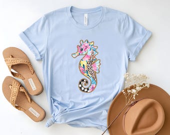 May include: Light blue t-shirt featuring a colorful seahorse design. The seahorse has floral, leopard print, and checkered patterns. The shirt is paired with brown sandals and a brown hat.