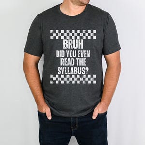 May include: Dark grey t-shirt with the text "BRUH DID YOU EVEN READ THE SYLLABUS?" in white, with a checkered pattern above and below the text. The shirt is worn by a person wearing dark blue jeans.