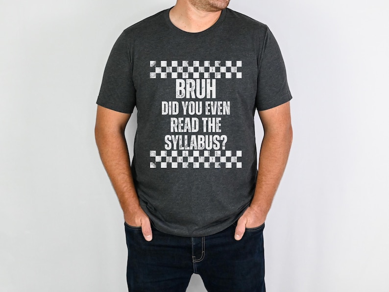 May include: Dark grey t-shirt with the text "BRUH DID YOU EVEN READ THE SYLLABUS?" in white, with a checkered pattern above and below the text. The shirt is worn by a person wearing dark blue jeans.