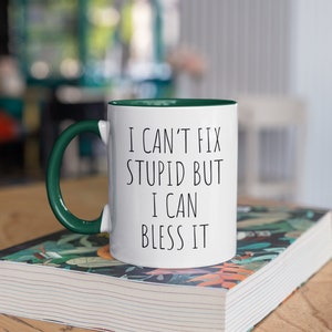 May include: White ceramic mug with a green rim and handle. The mug has black text that reads "I can't fix stupid but I can bless it". The mug is sitting on a book with a floral design.