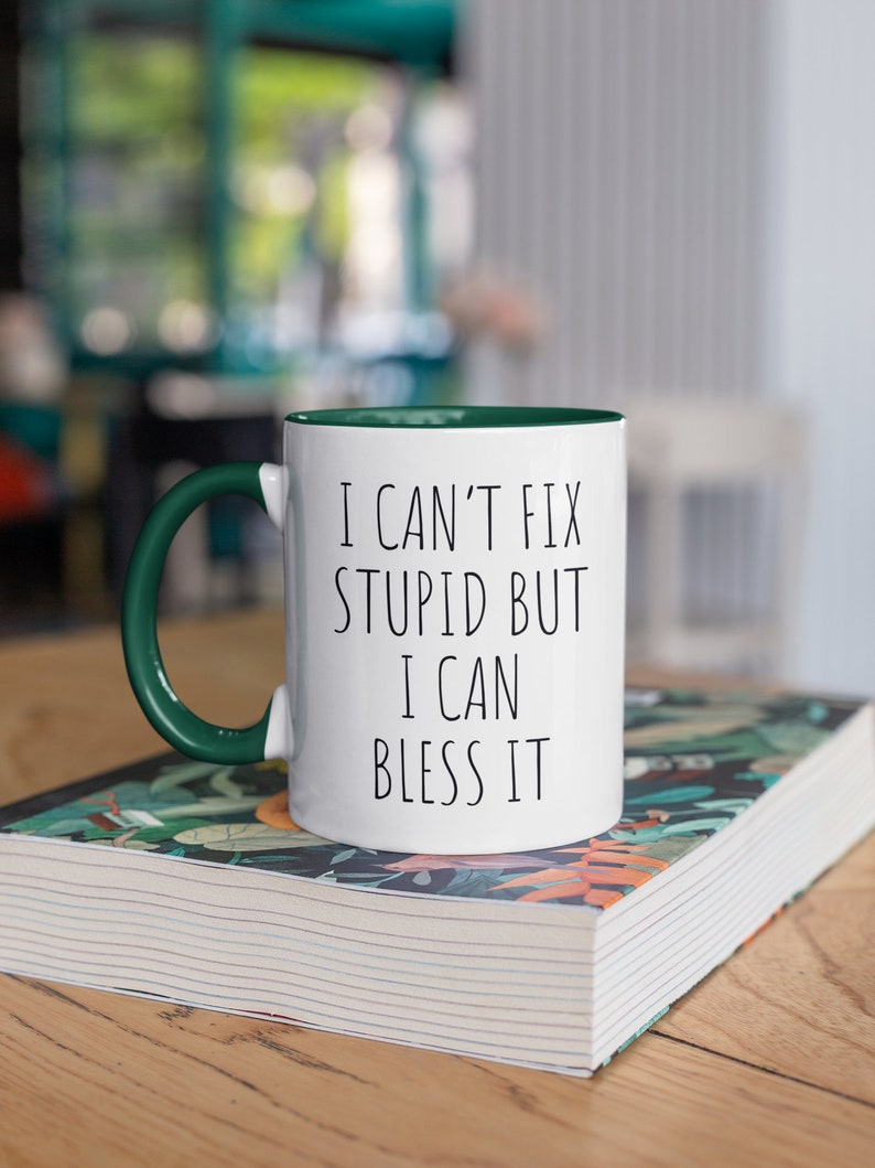 May include: White ceramic mug with a green rim and handle. The mug has black text that reads "I can't fix stupid but I can bless it". The mug is sitting on a book with a floral design.