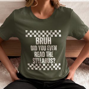 May include: An olive green t-shirt featuring the white text "BRUH DID YOU EVEN READ THE SYLLABUS?" with a checkered design above and below. The shirt has short sleeves.