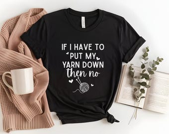 May include: Black t-shirt with the text "IF I HAVE TO PUT MY YARN DOWN THEN NO" in white. The design includes a yarn ball and knitting needles. A beige sweater, a white mug, and an open book are also in the image.