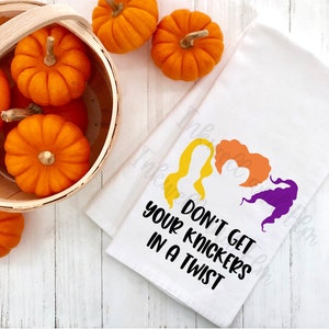 May include: White kitchen towel with a black graphic of the Sanderson sisters from the movie Hocus Pocus. The text reads "Don't get your knickers in a twist".  A basket of orange pumpkins is in the background.