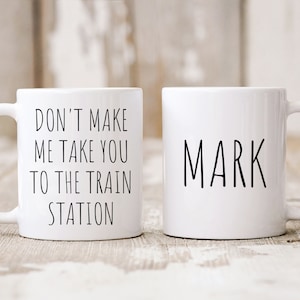 May include: Two white ceramic mugs with black text. One mug says "Don't make me take you to the train station" and the other mug says "Mark".