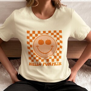 May include: A cream colored t-shirt with a checkered pattern and a smiley face with pumpkins for eyes. The text "HELLO PUMPKIN" is printed below the smiley face.