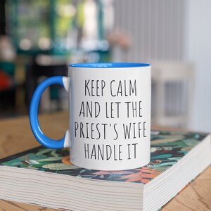 Peut inclure: Une tasse en c&eacute;ramique blanche avec un bord et une anse bleus. La tasse porte une inscription noire qui dit "Keep Calm and Let the Priest's Wife Handle It". La tasse est pos&eacute;e sur un livre &agrave; motifs floraux.