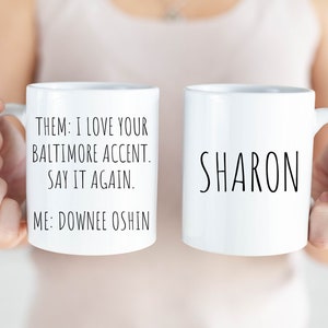 May include: Two white ceramic mugs. One mug has the text "THEM: I LOVE YOUR BALTIMORE ACCENT. SAY IT AGAIN. ME: DOWNEE OSHIN" and the other mug has the text "SHARON".
