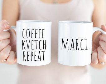 May include: Two white ceramic mugs. One mug says "Coffee Kvetch Repeat" and the other mug says "Marci".