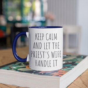Peut inclure: Une tasse en c&eacute;ramique blanche avec un bord et une anse bleus. La tasse porte une inscription noire qui dit "Keep Calm and Let the Priest's Wife Handle It". La tasse est pos&eacute;e sur un livre avec un motif floral.