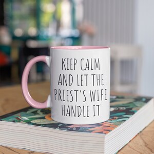 Peut inclure: Une tasse en c&eacute;ramique blanche avec un bord et une anse roses. La tasse porte une inscription noire qui dit "Keep Calm and Let the Priest's Wife Handle It". La tasse est pos&eacute;e sur un livre &agrave; motifs floraux.