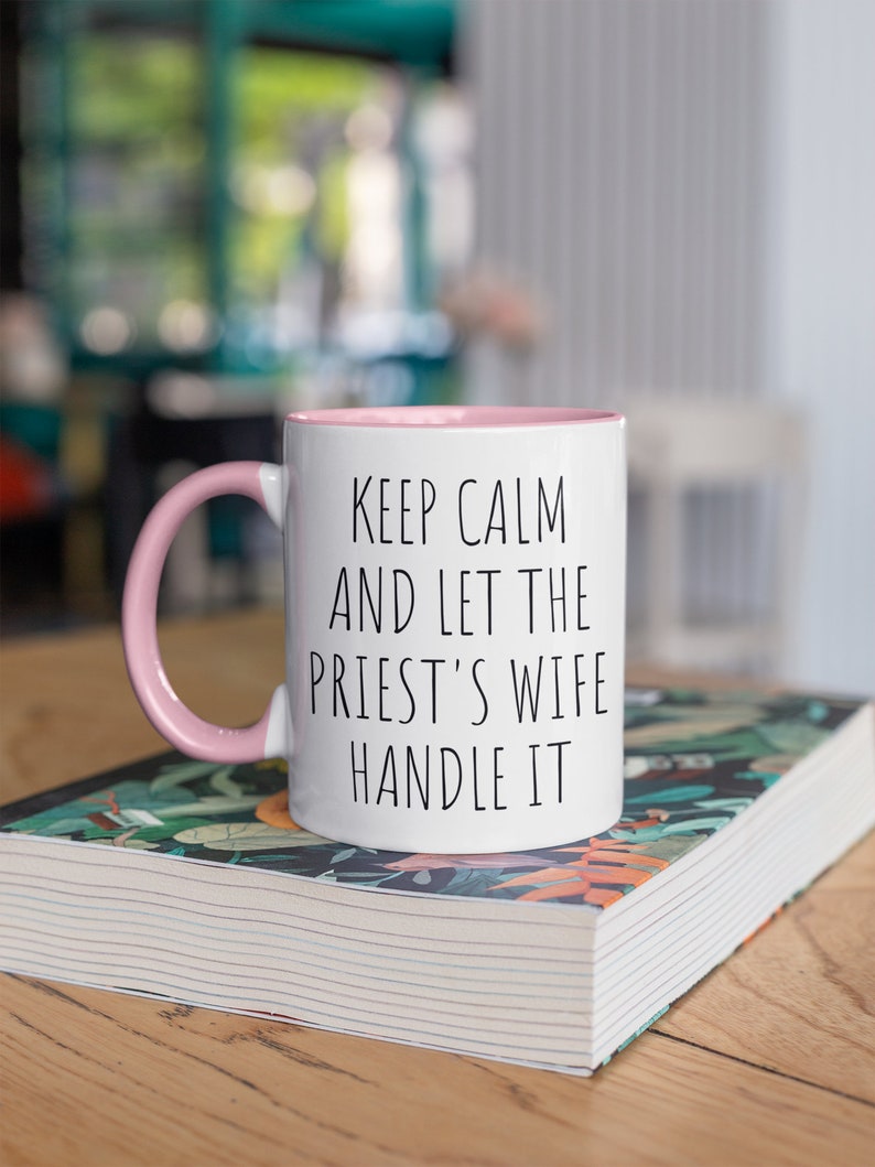 Peut inclure: Une tasse en c&eacute;ramique blanche avec un bord et une anse roses. La tasse porte une inscription noire qui dit "Keep Calm and Let the Priest's Wife Handle It". La tasse est pos&eacute;e sur un livre &agrave; motifs floraux.