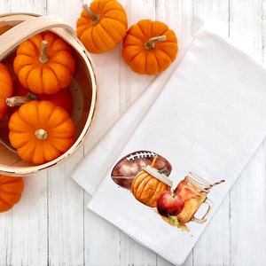 May include: A white kitchen towel with a watercolor illustration of a football, a pumpkin spice latte, and a red apple. The towel is surrounded by a wooden basket filled with orange pumpkins.