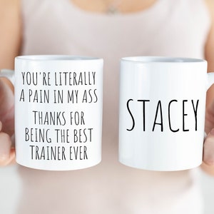 May include: Two white ceramic mugs. One mug has the text "YOU'RE LITERALLY A PAIN IN MY ASS THANKS FOR BEING THE BEST TRAINER EVER" in black. The other mug has the name "STACEY" in black. Both mugs have a white handle.