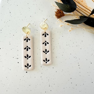 May include: A pair of earrings featuring a rectangular, off-white design with a series of black floral patterns. Each earring has a small, round, gold-colored stud at the top. The earrings are displayed on a speckled white surface.