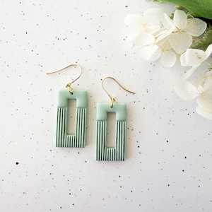 May include: A pair of rectangular earrings in shades of green, with a gold-colored hook. The earrings have a striped design on the lower half of the rectangle. The background is white with small black speckles, and white flowers are visible.