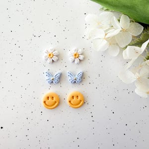 May include: A collection of decorative embellishments, including two white daisy flowers with yellow centers, two light blue butterfly shapes with black dots, and two yellow smiley face designs. The items are arranged on a white surface with black speckles, with white flowers and green leaves in the background.