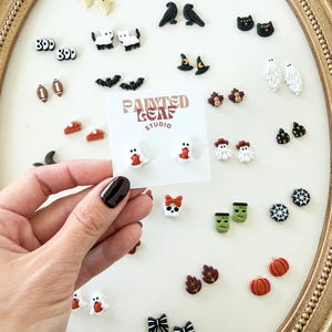 May include: A collection of small, colorful stud earrings featuring various Halloween and autumn themes, including ghosts, pumpkins, bats, skulls, spiders, and leaves. A white display card with the text Painted Leaf Studio is held in the center.
