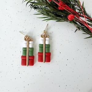 May include: A pair of festive earrings shaped like stacked gift boxes. Each earring features a red, green, and white color scheme, with gold-colored accents. The earrings are set against a white background with greenery and red berries.