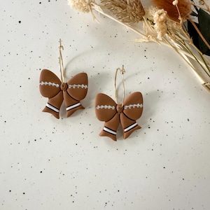 May include: A pair of brown bow-shaped earrings with white and black accents. The earrings are on gold hoops. The background is a speckled white surface with dried flowers.