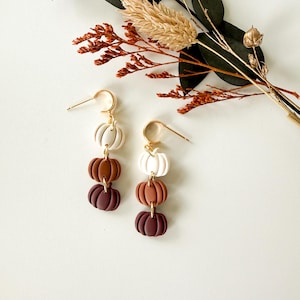 May include: A pair of drop earrings featuring three stacked pumpkin charms in graduated shades of cream, terracotta, and dark brown. The charms are suspended from gold-toned metal hoops. Each pumpkin has a ribbed texture and a rounded, segmented shape.