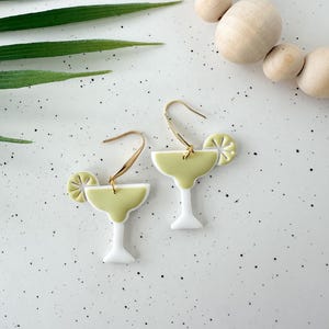 May include: A pair of gold dangle earrings with a white and green margarita glass charm and a lime slice. The earrings are on a white speckled surface.