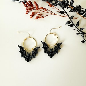 May include: A pair of dangle earrings featuring gold-toned circular hoops attached to matte black bat-shaped charms. The bat charms display intricate, symmetrical embossed scrollwork patterns across their wings and bodies, suspended from simple gold-toned ear wires.