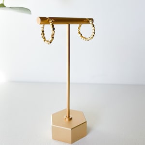 May include: Gold hoop earrings with a textured, geometric design. The earrings are displayed on a gold stand.
