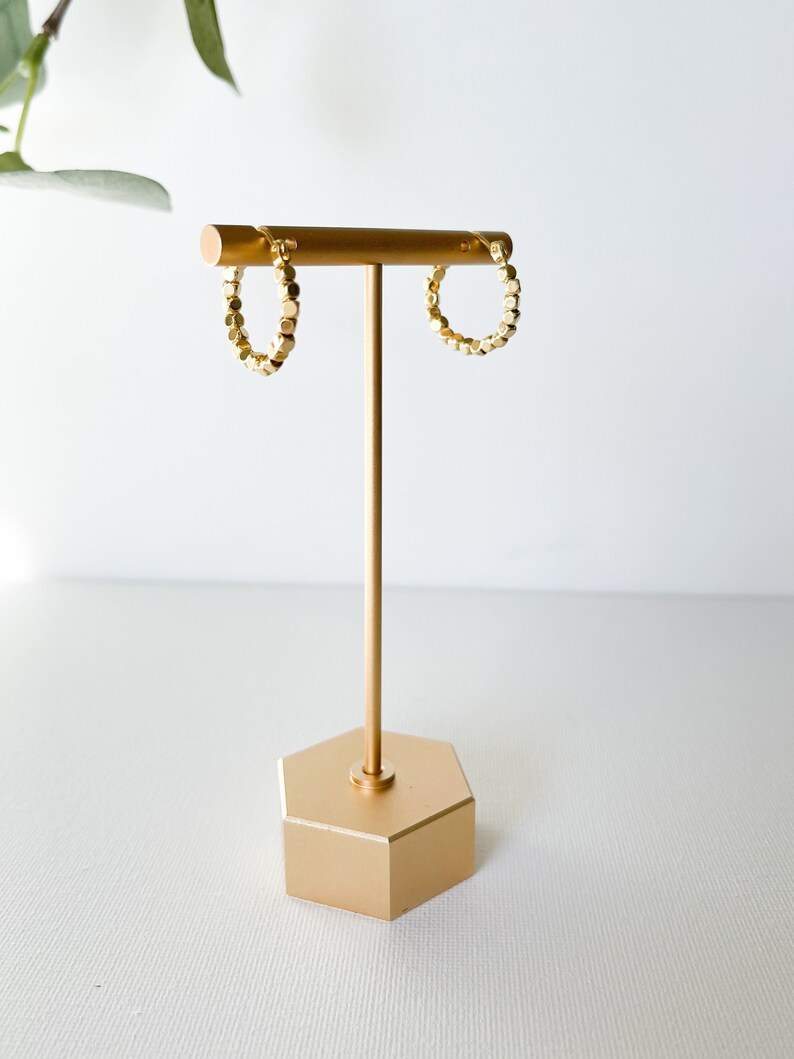 May include: Gold hoop earrings with a textured, geometric design. The earrings are displayed on a gold stand.
