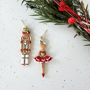 May include: Gold-toned dangle earrings featuring a Christmas theme. One earring is a nutcracker soldier in cream, gold, and brown. The other is a ballerina in a red tutu. Greenery and red berries are in the background.