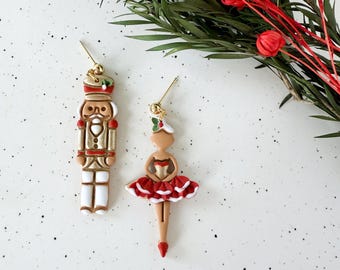 May include: Gold-toned dangle earrings featuring a Christmas theme. One earring is a nutcracker soldier in cream, gold, and brown. The other is a ballerina in a red tutu. Greenery and red berries are in the background.